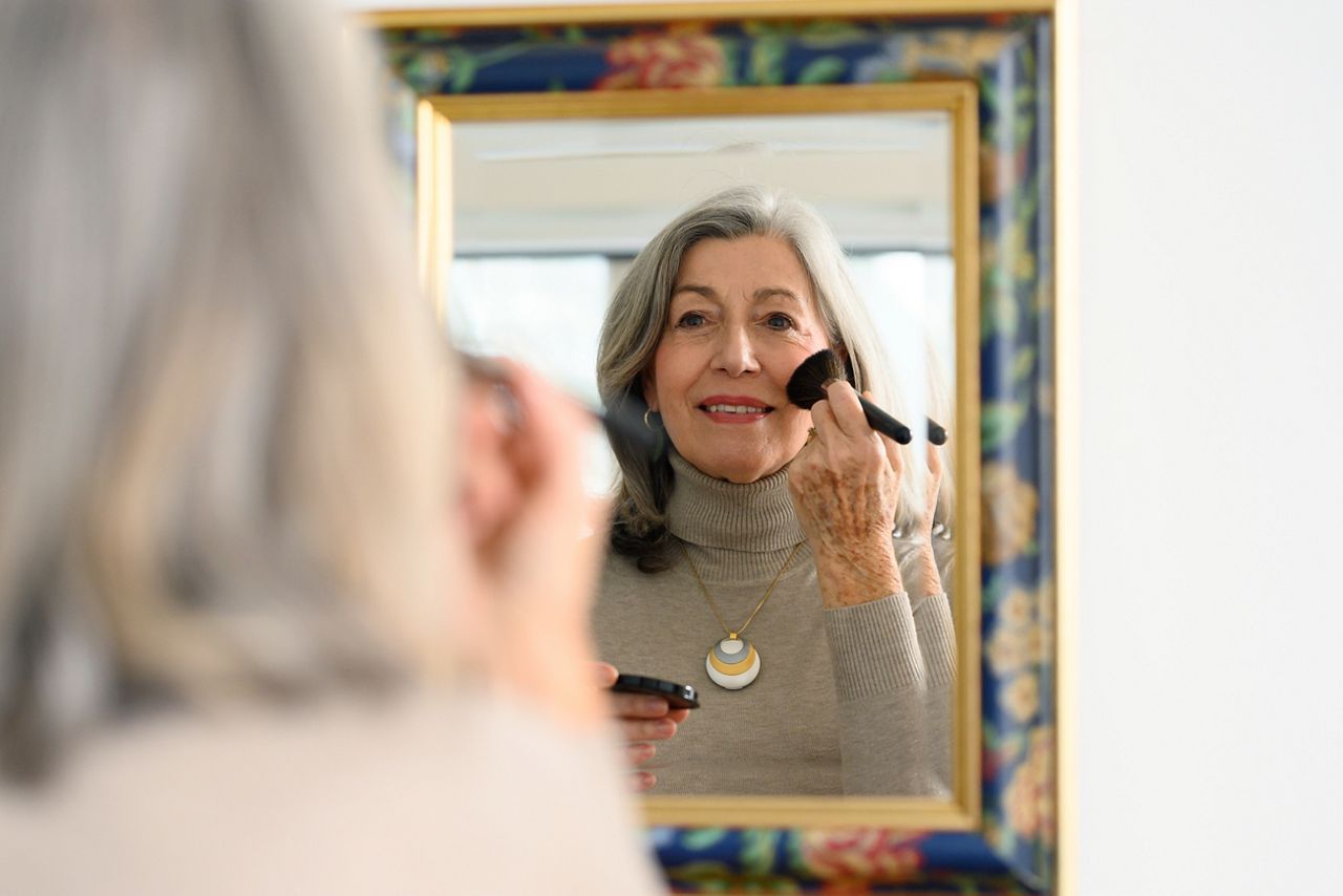 Oudere vrouw met een persoonlijk alarmhangertje kijkt in de spiegel terwijl ze haar make-up opdoet.