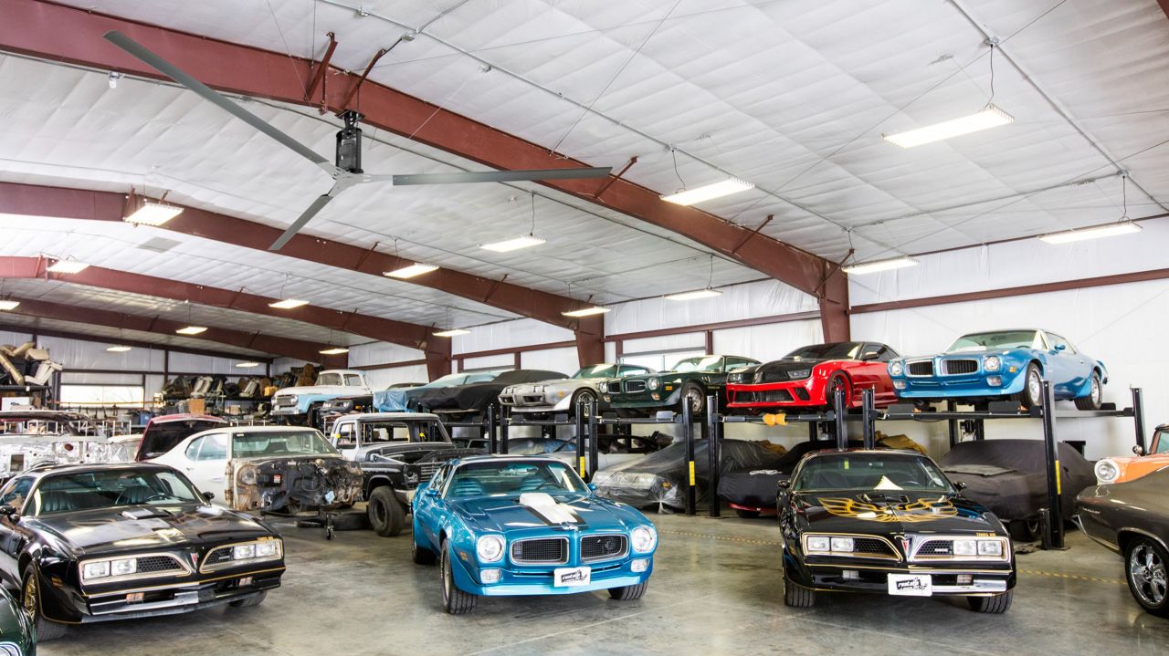 A black ceiling fan with three blades hanging in a car shop above multiple vintage cars