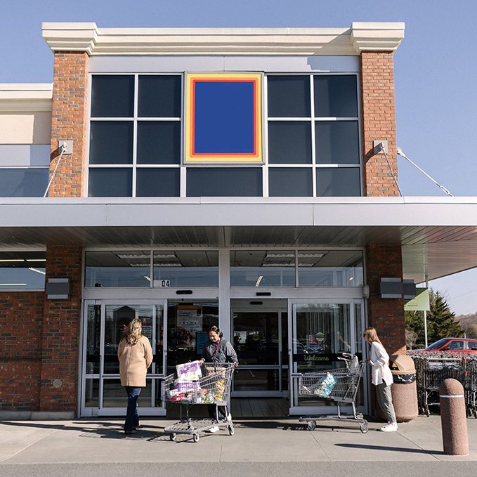 Image: Front entrance of a grocery store with brick and gray exterior, large glass windows, and sliding glass doors. Several individuals stand near shopping carts on the sidewalk in front of the entrance.