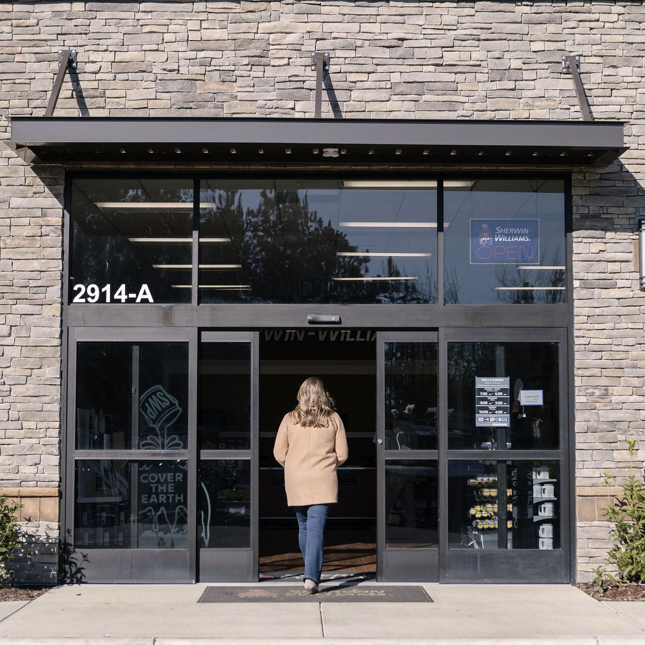 Image: Exterior view of a stone-front retail store with large glass double doors. A person wearing a tan coat and jeans is walking into the open automatic door.