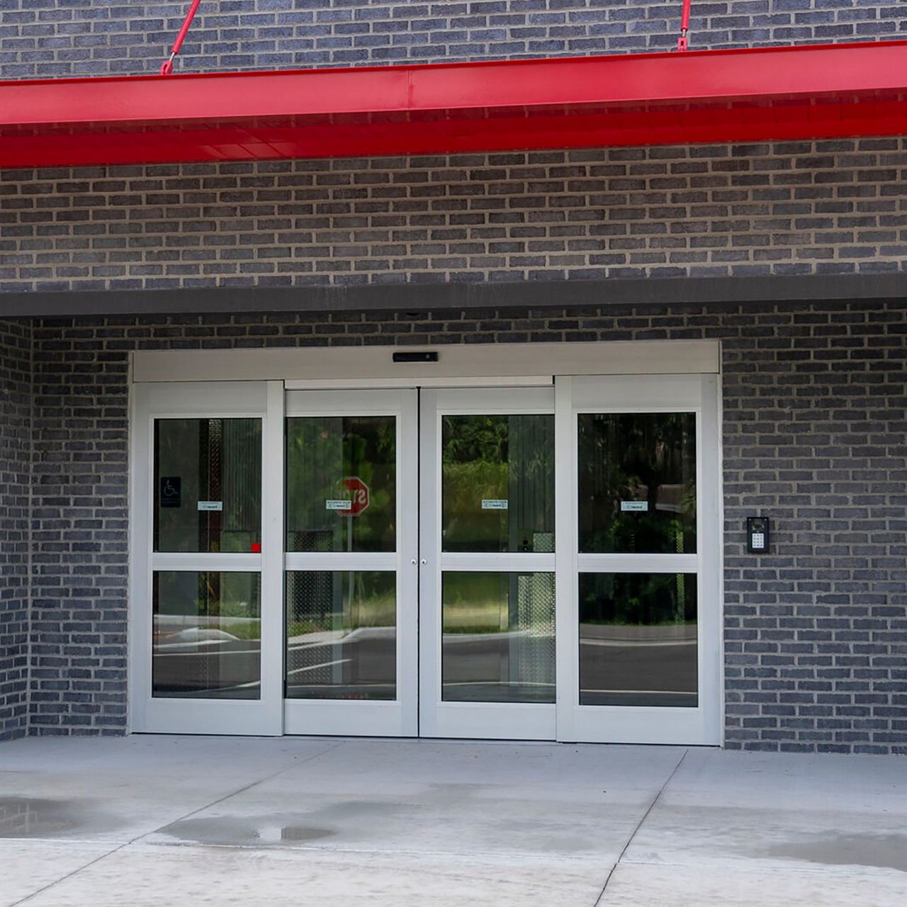 A clear, bi-parting automatic door on a brick building with a red overhang.