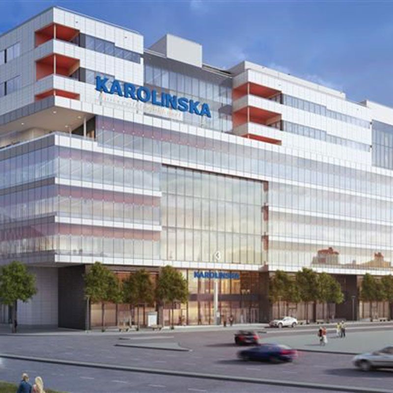 Visualisering framifrån av Nya Karolinska Sjukhusbyggnaden. En stor vit byggnad med många stora glasfönster och dörrar. Framför byggnaden finns bilar som kör och människor som går. 
