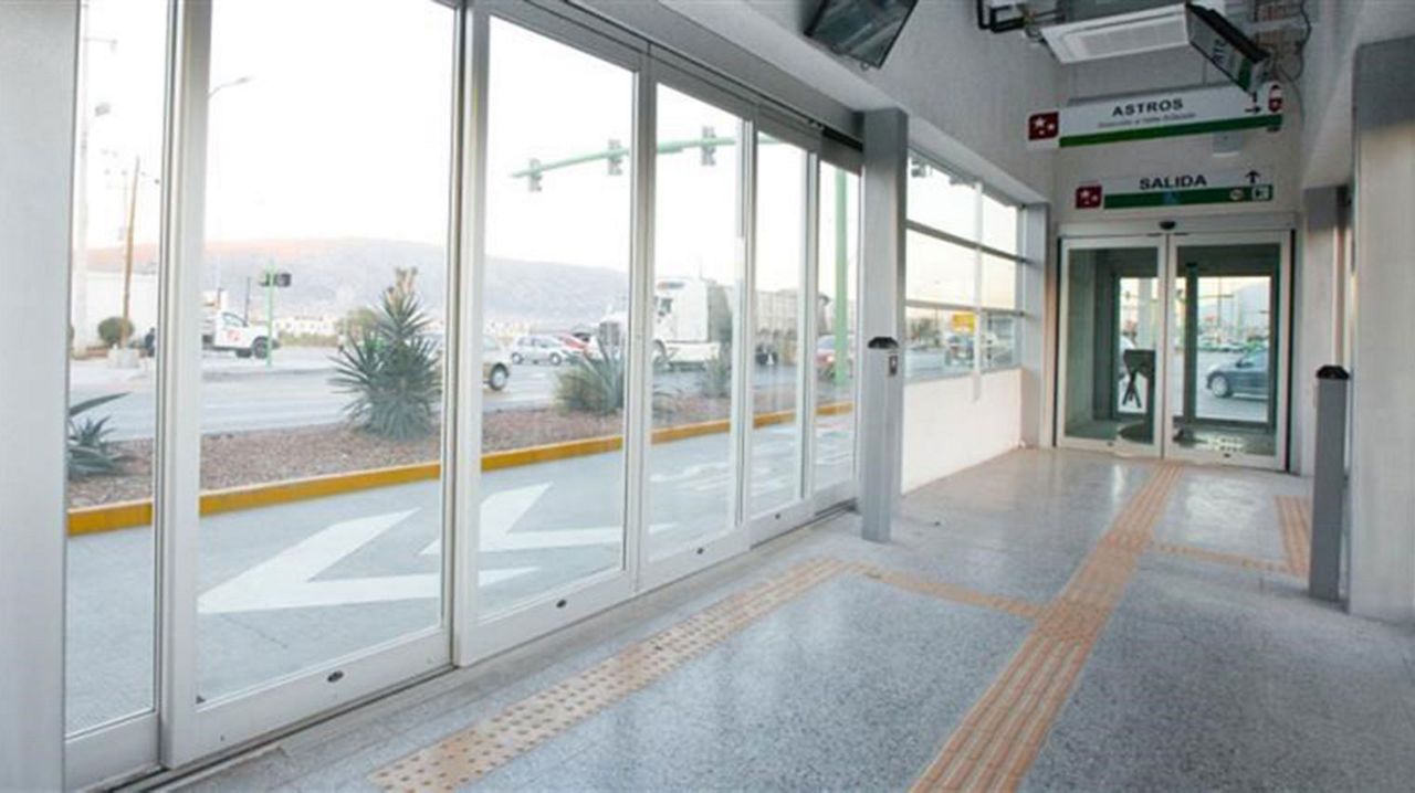 Transit hall with automatic door systems at the Ecovía BRT system. Big glass windows and doors. Outside you can see small palm trees and a sidewalk.