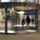 Two persons walking through revolving doors. The door has arrows on it and on the side of the doors there are pillars and trashcans. 