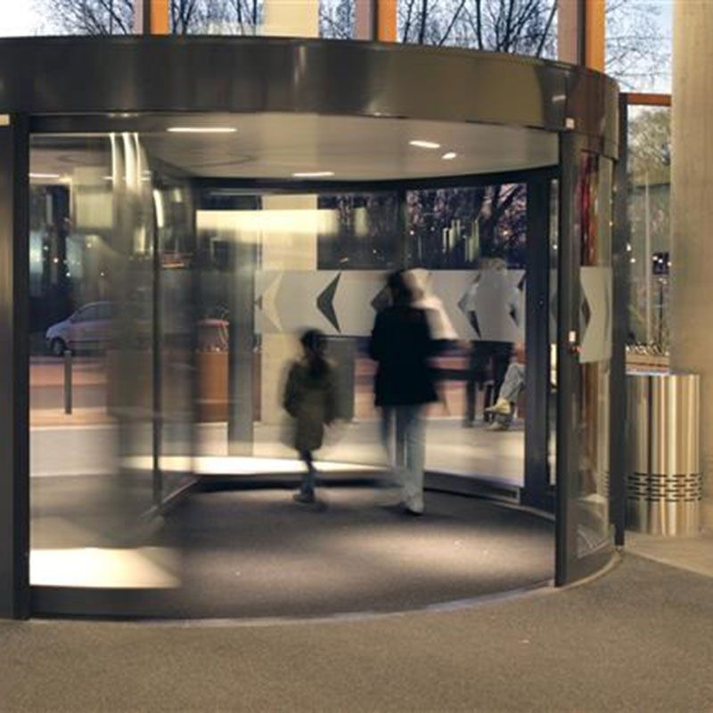 Two persons walking through revolving doors. The door has arrows on it and on the side of the doors there are pillars and trashcans. 