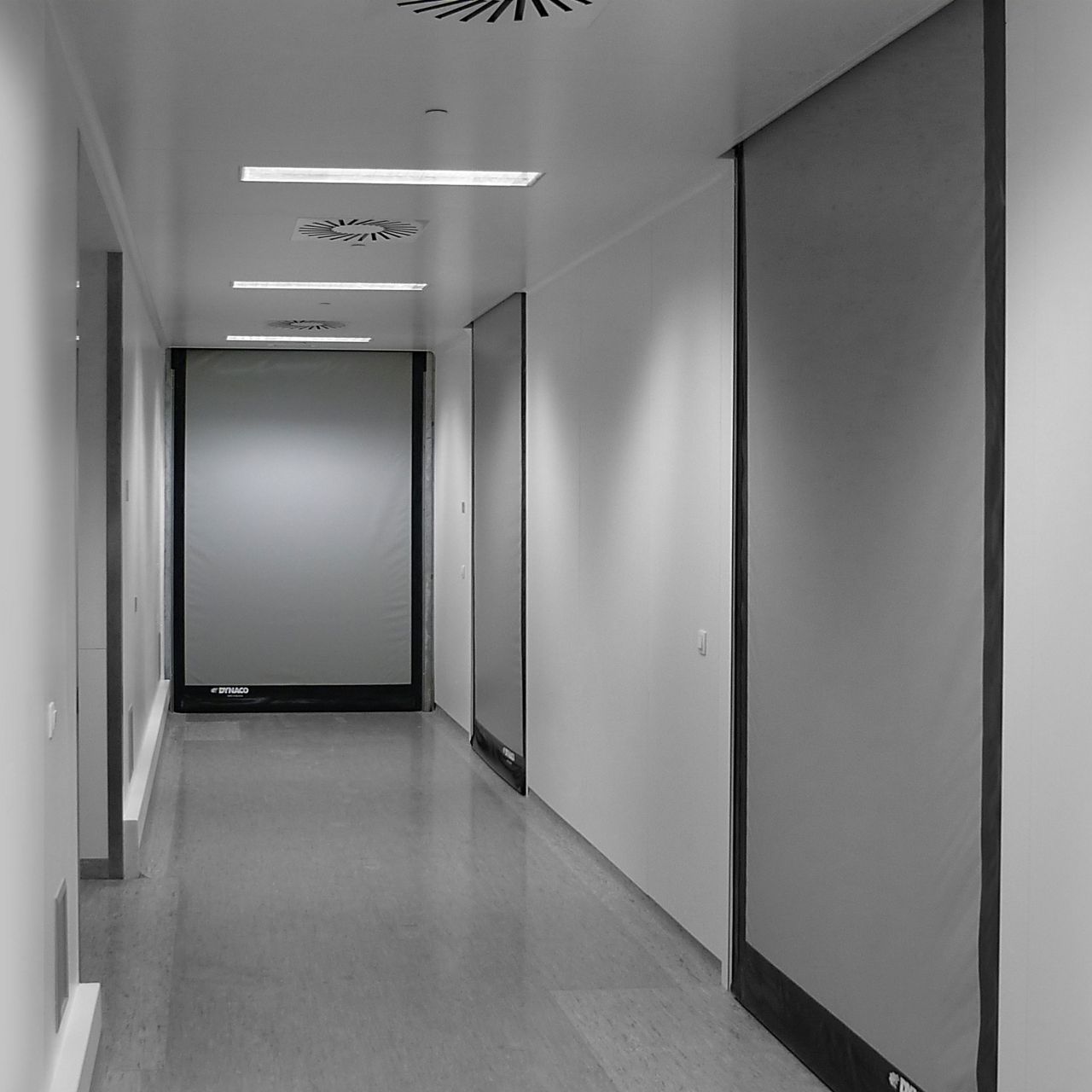 A grey corridor and grey doors that are open, and you can see a person in the background. 