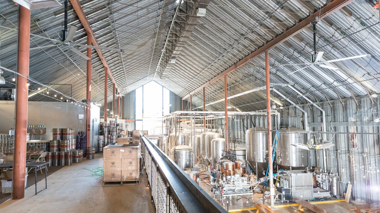 Two silver fans with 5 blades that have curved, black edges hanging in a brewery over a conveyor belt