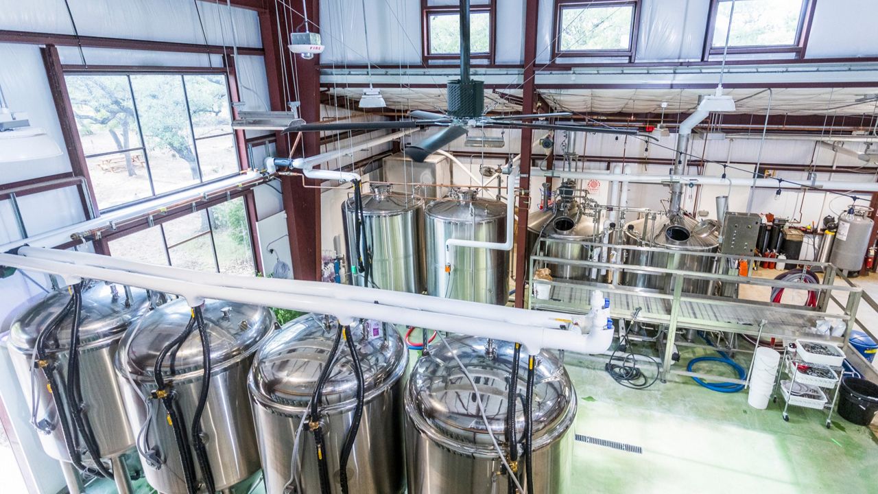 A silver fan with 5 blades that have curved, black edges hanging over barrels of beer in a brewery