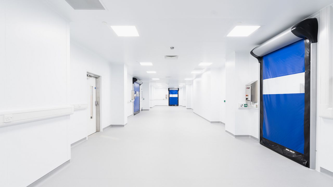 Image: Porta Rápida para Sala Limpa ASSA ABLOY HS9130GAT, azul com visor horizontal transparente, instalada em corredor de ambiente controlado com múltiplas áreas limpas para processos farmacêuticos, laboratoriais ou industriais.