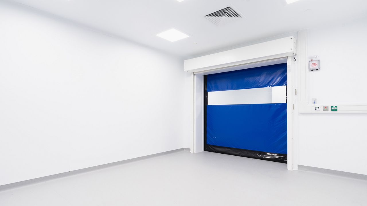 Image: Porta Rápida para Sala Limpa ASSA ABLOY HS9130GAT, azul com visor horizontal transparente, instalada em ambiente estéril de área limpa para controle de acesso, separação de áreas e manutenção das condições de higiene e contaminação controlada.