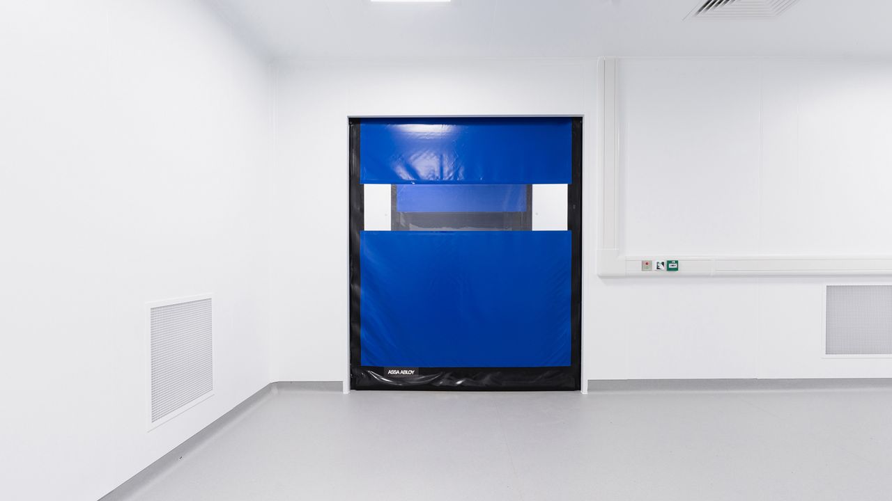 Image: Porta Rápida para Sala Limpa ASSA ABLOY HS9130GAT, azul com visor horizontal transparente, instalada em ambiente estéril de área limpa para controle de acesso, separação de áreas e manutenção das condições de higiene e contaminação controlada.
