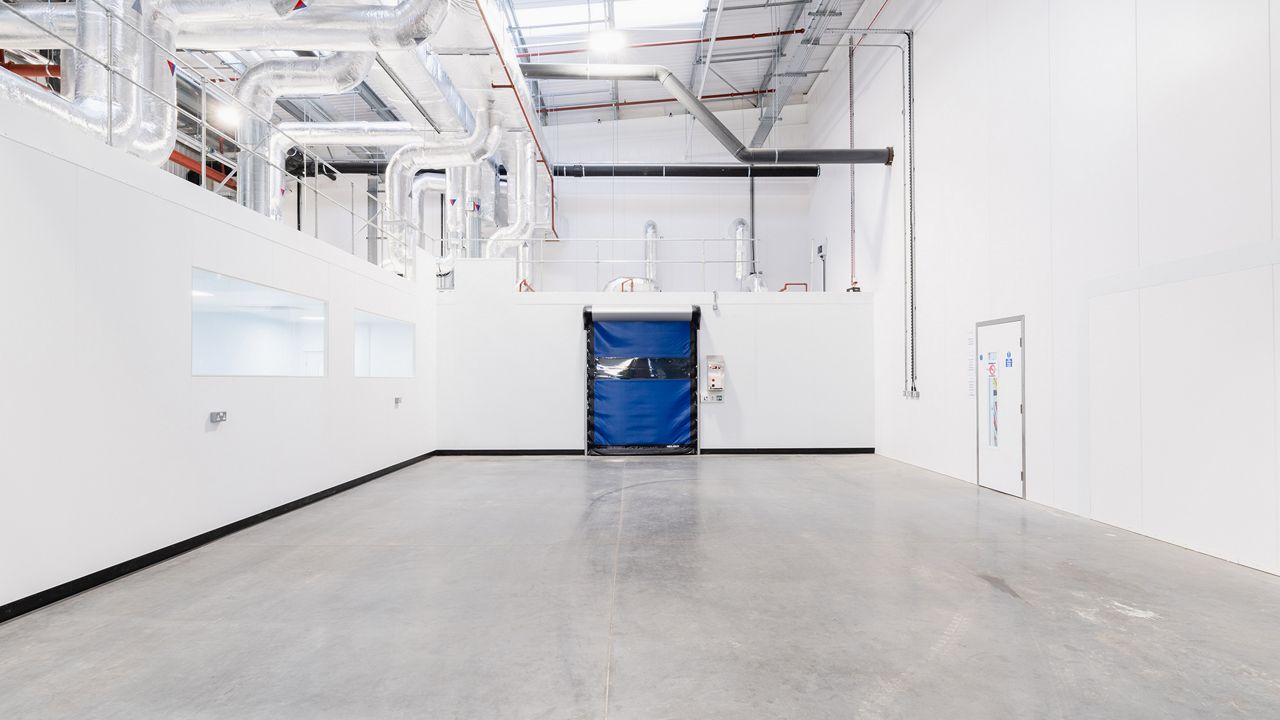 Image: Porta Rápida para Sala Limpa ASSA ABLOY HS9130GAT, azul com visor transparente horizontal, instalada em ambiente industrial de produção com áreas limpas e infraestrutura HVAC aparente para controle de contaminação e circulação de ar.