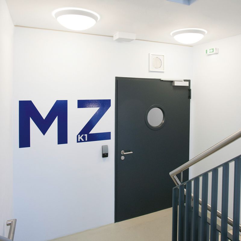 Schwarze Tür mit rundem Fenster in einem Treppenhaus mit der Bezeichnung "MZ" daneben