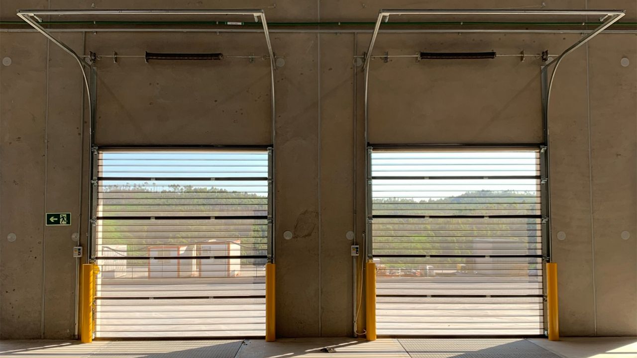 Image: Porta Seccional Transvision Inovadoor Série 400, com painéis transparentes de alta visibilidade, instalada em docas de carregamento de centro logístico