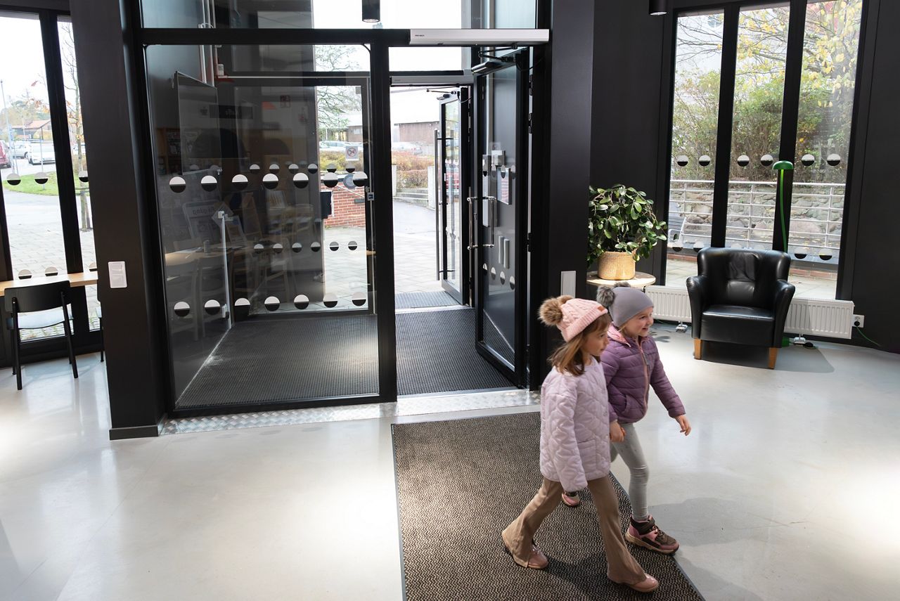 Prosklené zádveří s prosklenými dveřmi s dveřním pohonem ASSA ABLOY SW300-S v popředí jsou procházející děti