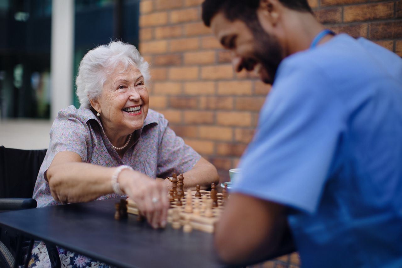 Senior woman and male nurse sitting at a table outdoors playing chess.