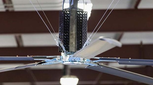 A fan with 5 black blades and a silver center hanging in a warehouse over multiple vintage cars