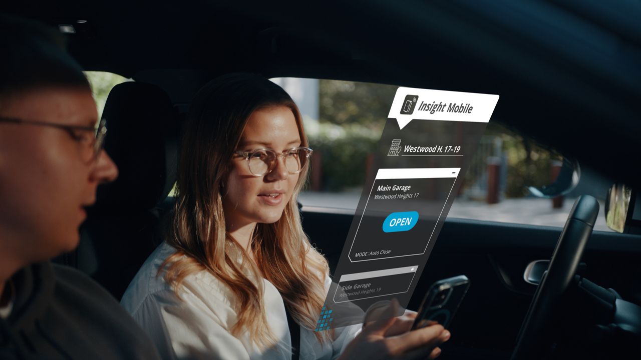Žena a muž v autě, žena drží telefon se zobrazenou aplikací Insight Mobile a tlačítkem „Otevřít“ pro hlavní garážová vrata.