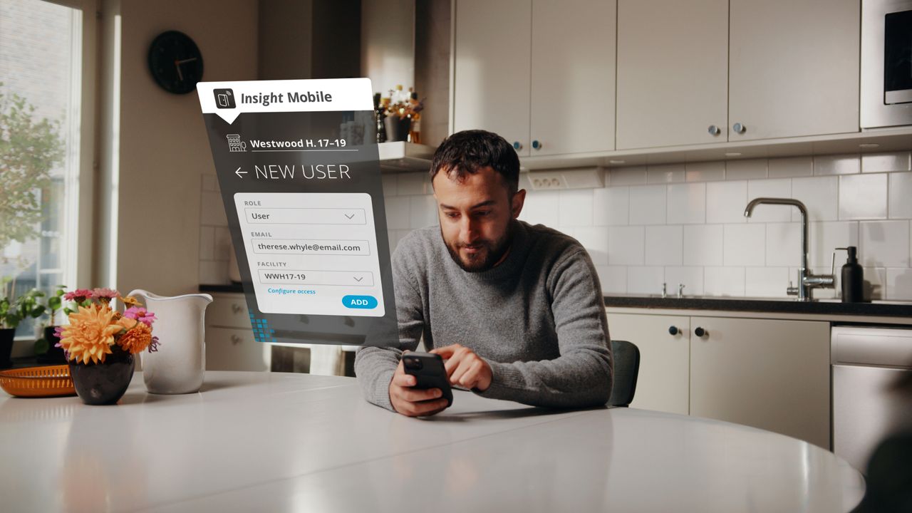 Man sitting at a table in a kitchen, holding a phone displaying the Insight Mobile app interface where he appears to be adding a new user to a location
