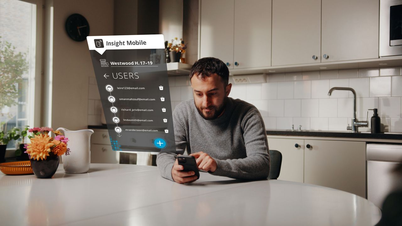 Man sitting at a table in a kitchen, holding a phone displaying the Insight Mobile app interface with a list of email addresses under a 'Users' tab