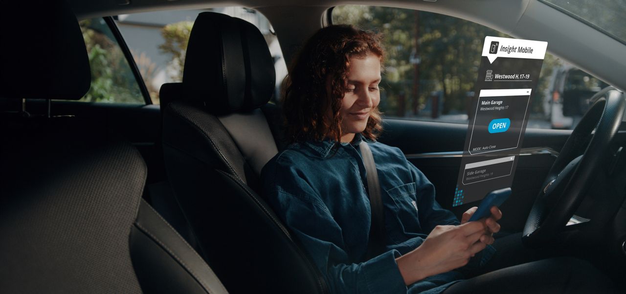 Woman in a car, holding a phone displaying the Insight Mobile app interface with an 'Open' button for a main garage door