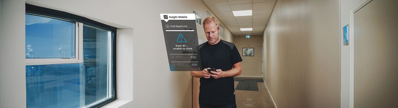 Man in a corridor of a building, holding a phone displaying the Insight Mobile app interface with a notification that says 'door unable to close'