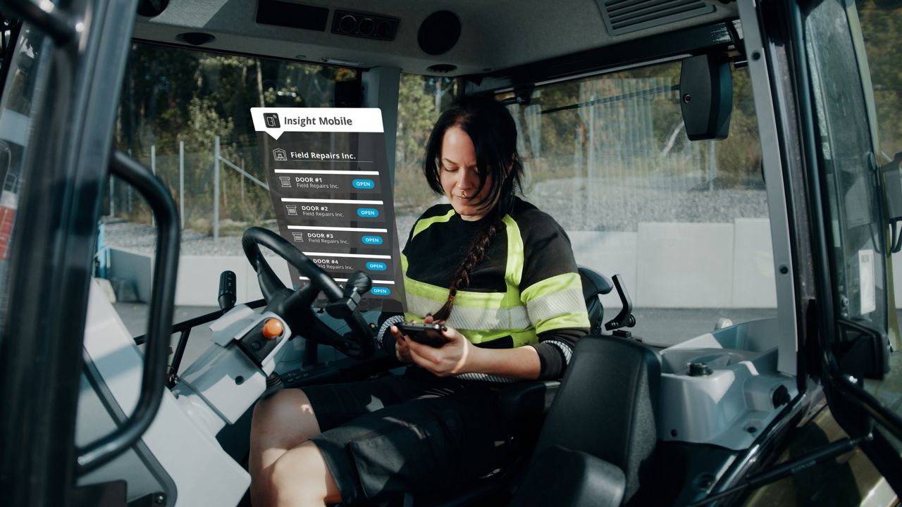 Woman inside a tractor, holding a phone displaying the Insight Mobile app interface with a list of doors and 'Open' buttons next to each door