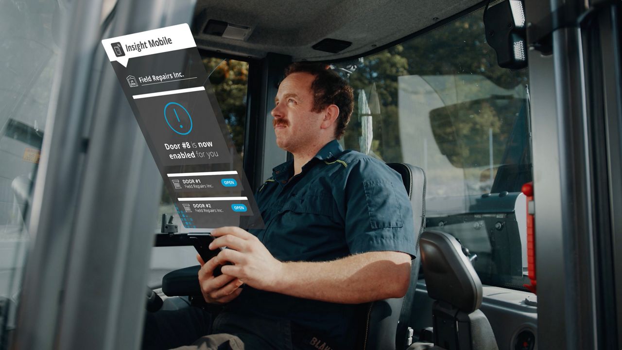Man inside a tractor, holding a phone displaying the Insight Mobile app interface that shows a 'Door enabled' notification and a list of doors and 'Open' buttons next to each door