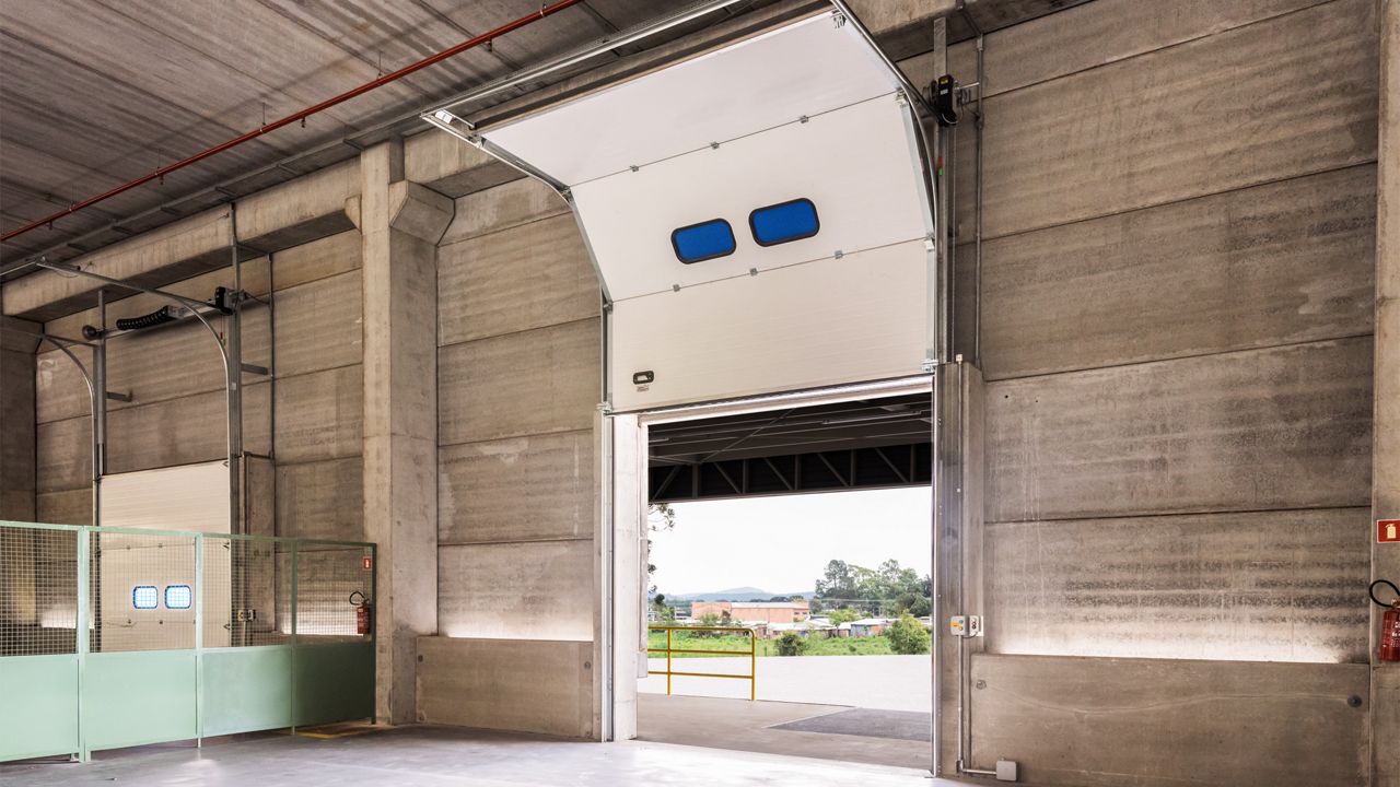 Image: Porta Seccional Industrial Inovadoor, branca com visores retangulares, instalada em centro logístico ou armazém industrial, aberta para permitir a movimentação eficiente de cargas, veículos e operações de doca.