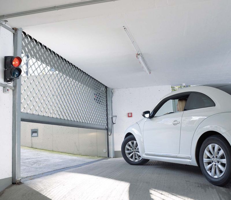Ein Auto steht vor einem Rollgitter, welches sich öffnet. Links an der Wand ist eine rote Ampel.