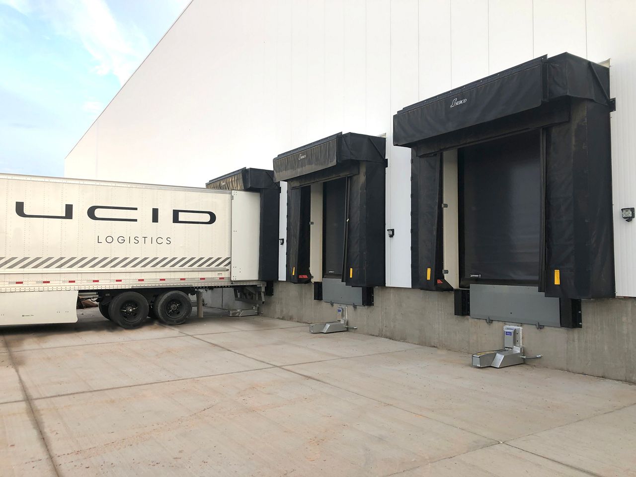 Image: 3 doors outside of a warehouse with black hinge guards. A white truck is backing into one of the doors to prepare for loading.