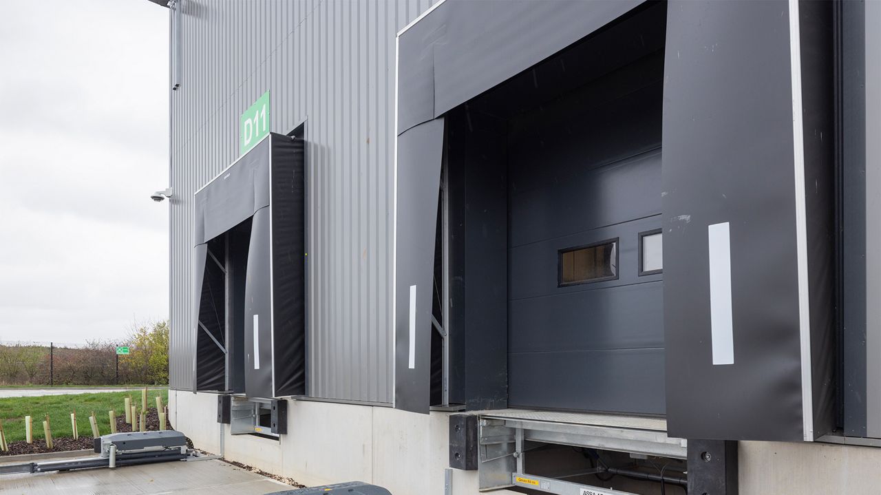Image: Abrigo de Doca Inovadoor 202, instalado em docas de carregamento de centro logístico, com cortinas de vedação frontal em lona resistente e faixas refletivas laterais de alta visibilidade