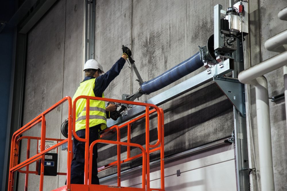 Porta industrial instalada em ambiente industrial, com técnico realizando manutenção no sistema de acionamento superior utilizando plataforma elevatória para inspeção e ajuste do equipamento.