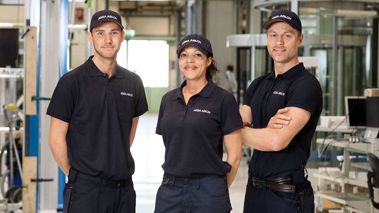 Três pessoas vestidas com roupa de trabalho Assa Abloy numa fila a sorrir para a câmara.
