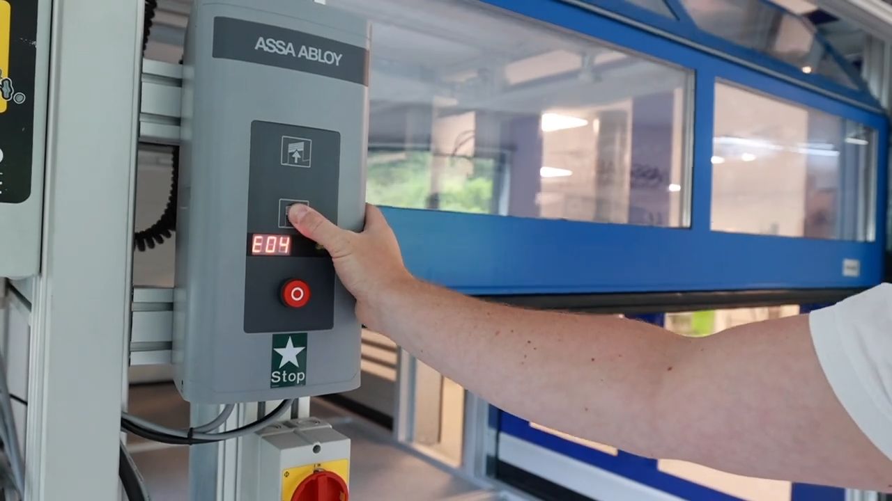 Controlepaneel van een ASSA ABLOY Crawford sectionale poort met foutcode E04 op het display. Een hand drukt op de sluitknop, maar de poort blijft hangen.