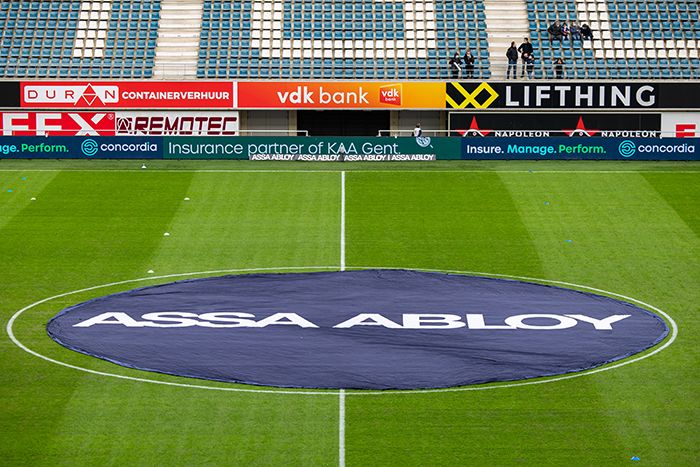 Uitzicht op het voetbalstadion Planet Group Arena van KAA Gent vlak voor de aftrap van een wedstrijd. Op de middenstip ligt een ronde banner met het logo van ASSA ABLOY duidelijk zichtbaar op het grasveld.