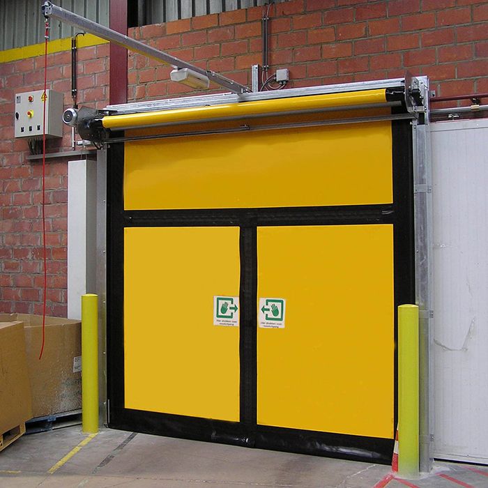 Porte rapide jaune à 3 panneaux dans un bâtiment industriel ; 2 panneaux sont des portes de secours et portent des symboles verts indiquant « Appuyer ici pour la sortie de secours ».