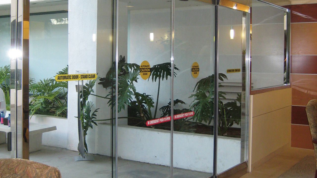 Une porte coulissante vitrée dans un hall avec des plantes vertes à l’extérieur. 