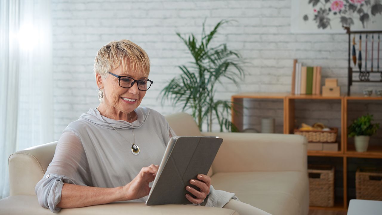 Lachende vrouw met een persoonlijk alarmhangertje gebruikt haar tablet terwijl ze op een bank zit.