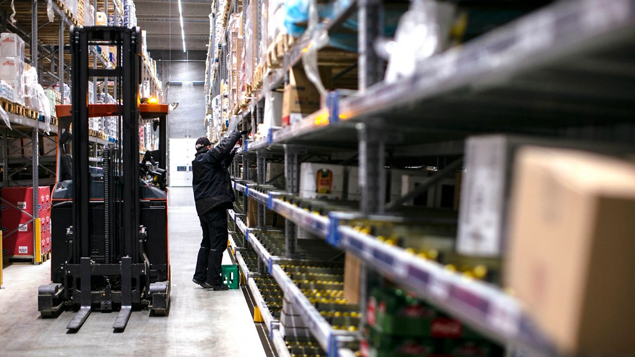 En mann som jobber i et stort lager står ved siden av gaffeltrucken sin og sjekker varer i hyllene.