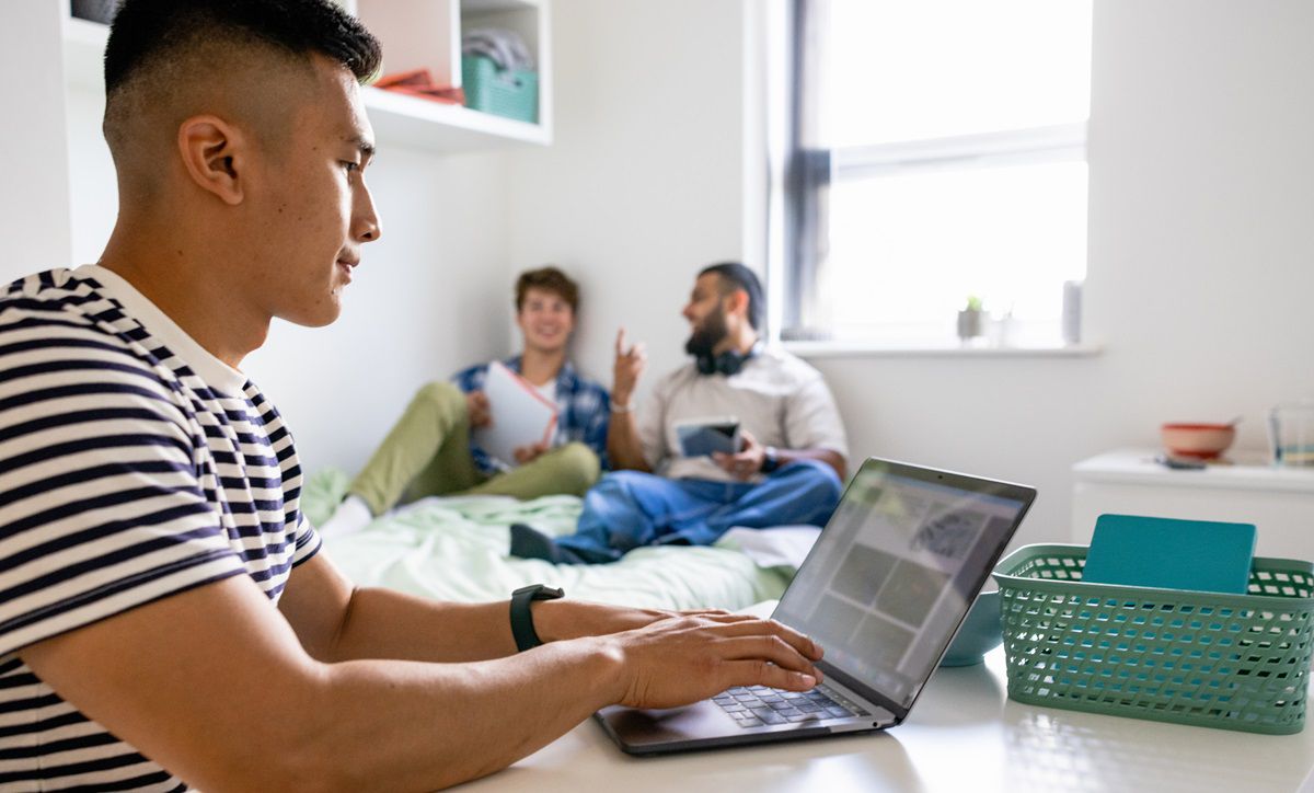 A small group of male uni student friends sitting in their dorm bedroom are studying together while sitting on the bed, one man is sitting at his desk.