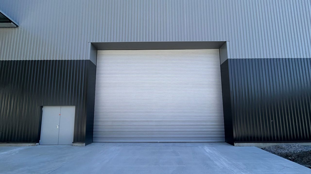 Dark grey industrial building with insulated metal roller shutter and small entrance door next to it, front view.