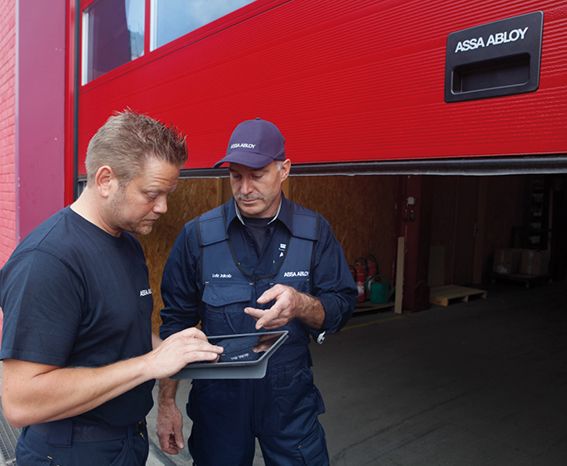 Twee mannen in ASSA ABLOY-werkkleding bekijken een tablet voor een open rode garagedeur van een industrieel gebouw.