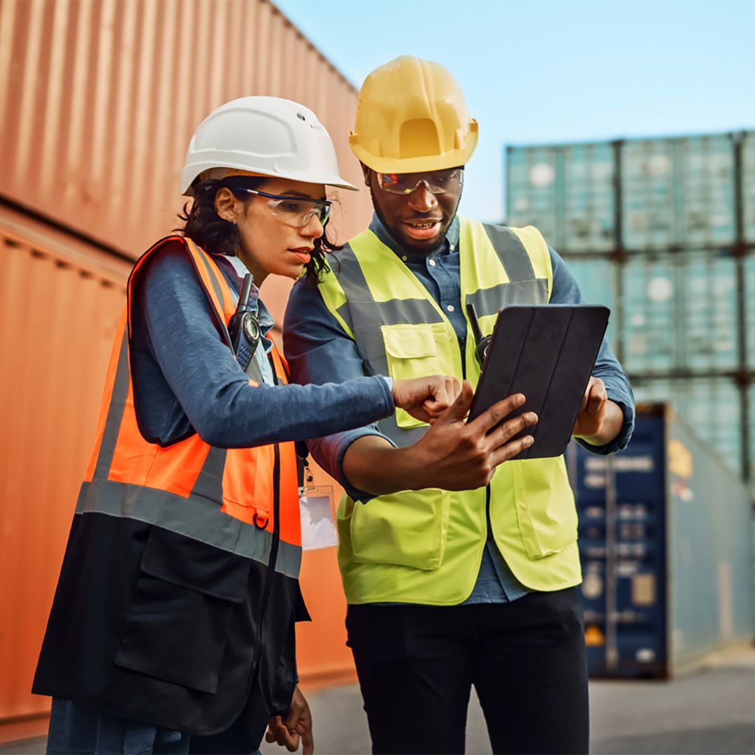Två personer står i en hamnmiljö och läser på en surfplatta.