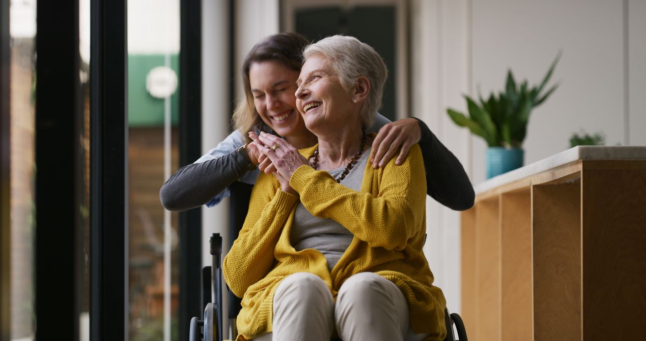 Female nurse higging senior woman in wheelchair at care home.