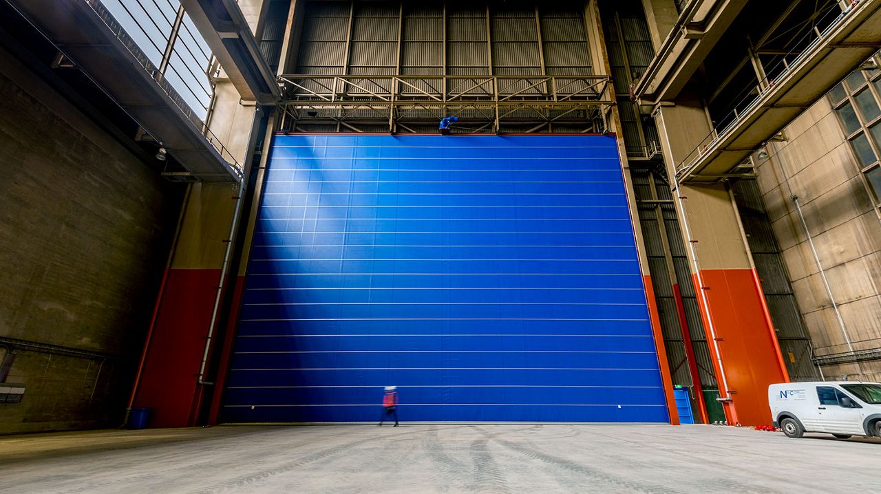 A big blue high speed door, a worker walking infront of it and on the left side you can see a white car. 