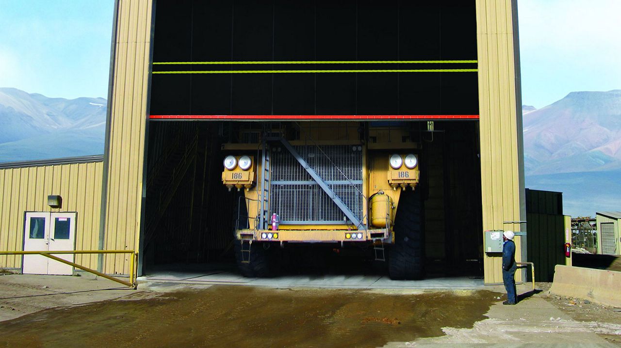 A big high speed door opening and you can see part of a large yellow truck. The building is pale yellow and the door is black. A worker is standing next to the opening. 