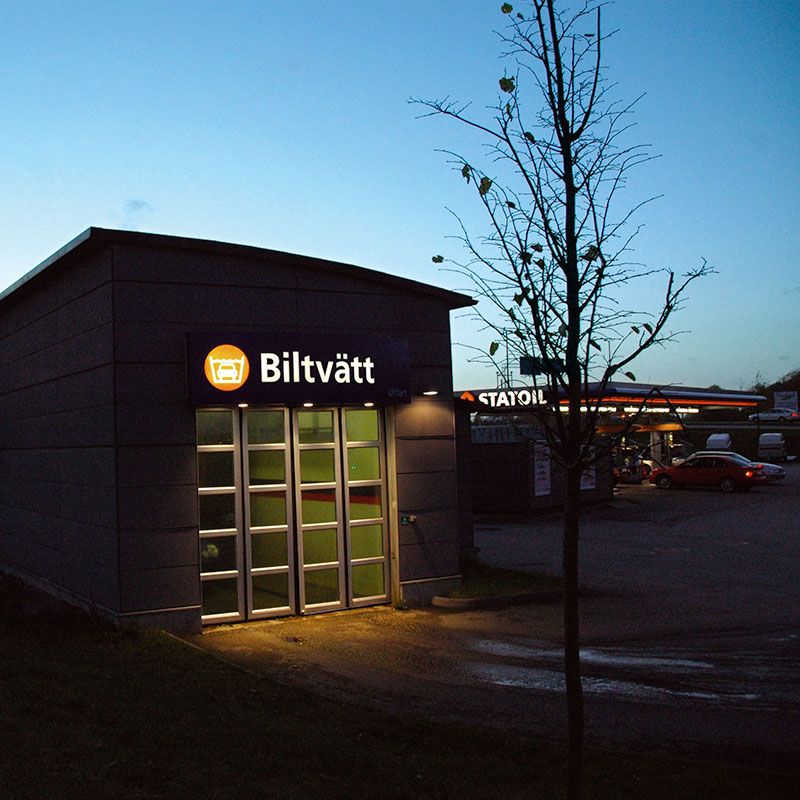 A picture of a car wash in the night. The car wash has glass doors and a orange sign, there is a tree infront of the car wash. 