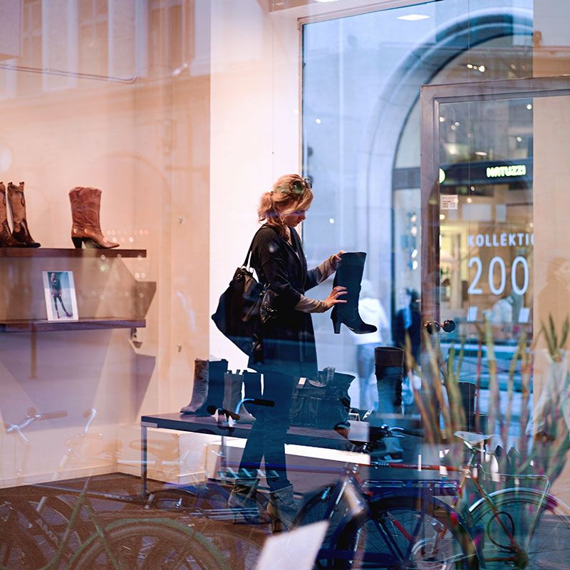 A woman looking at shoes in a fashion store. There are big windows and shoes and boots on display. 