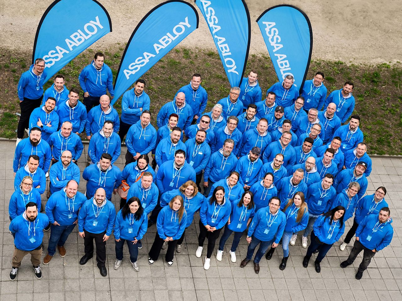 Mitarbeiter von ASSA ABLOY Automatiktüren in blauen Hoodies schauen Richtung Himmel. Im Hintergrund stehen vier blaue Beachflags mit ASSA ABLOY-Logo